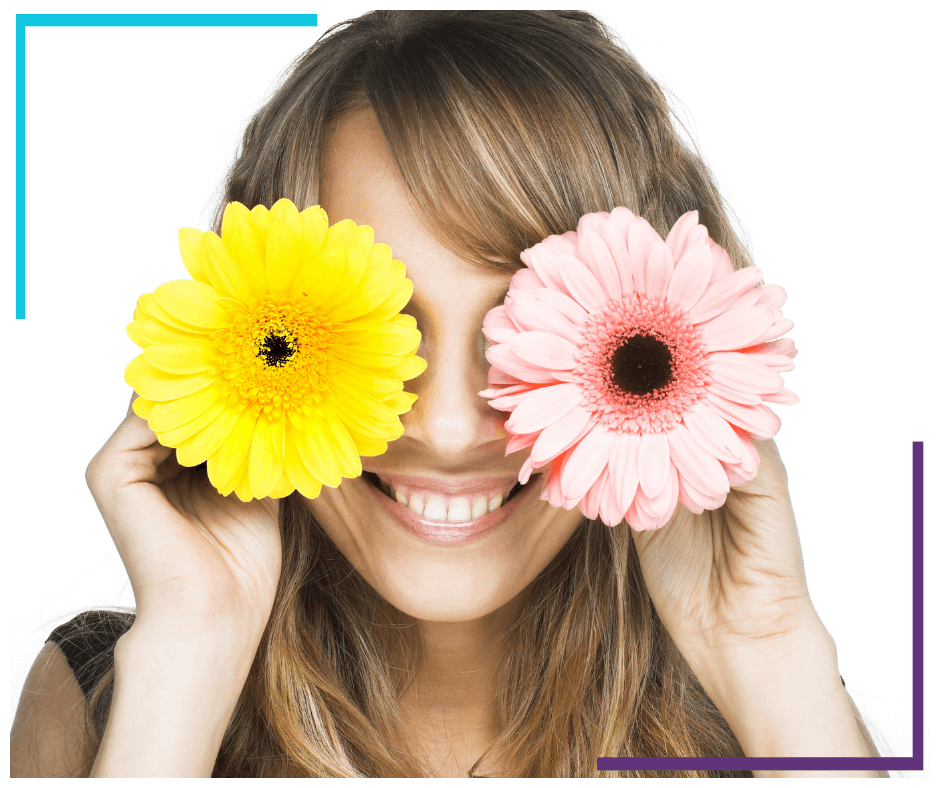 A woman holds a pink flower over her left eye and a yellow flower over her right eye. She's smiling.
