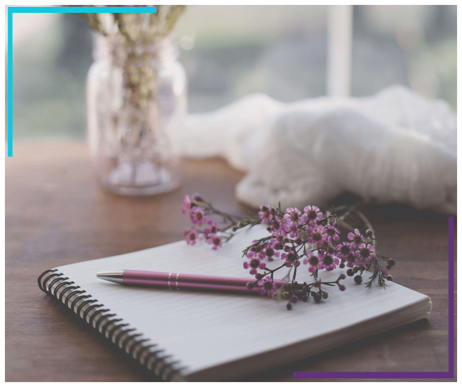 A journal sits open on a table, with purple flowers and a pen on top. In the background there's a vase with purple flowers in it and a white blanket that sits bundled on the edge of the table.