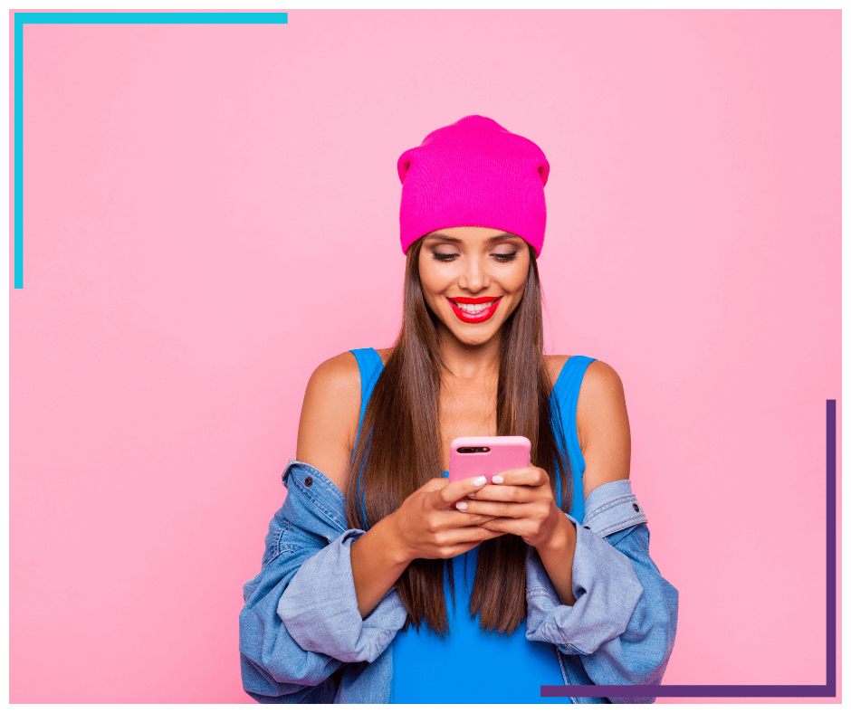 A woman stands in from of a pink backdrop, wearing a pink tuque. She looks down at the phone she's holding and smiles.