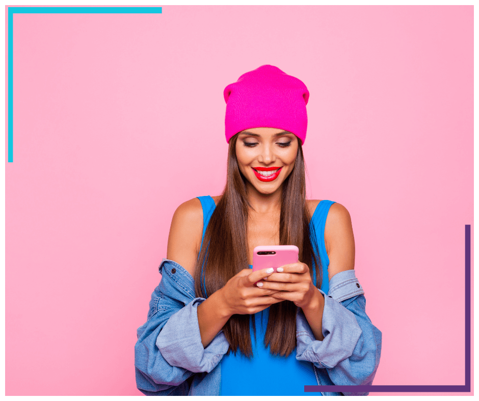 Photo of a woman wearing a jean jacket and pink tuque holding and looking at her cell phone. The background is also bright pink